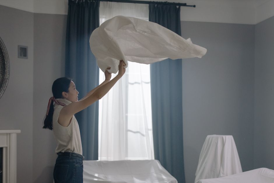 An Asian woman in a sleeveless shirt shakes out a white sheet in a well-lit room.